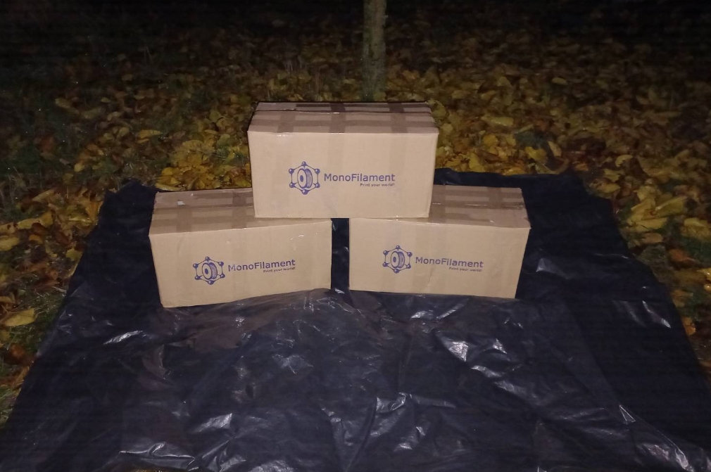 Три картонні коробки з логотипом MonoFilament стоять на чорному тенті серед опалого листя. Коробки містять пластик PLA для 3D-друку.